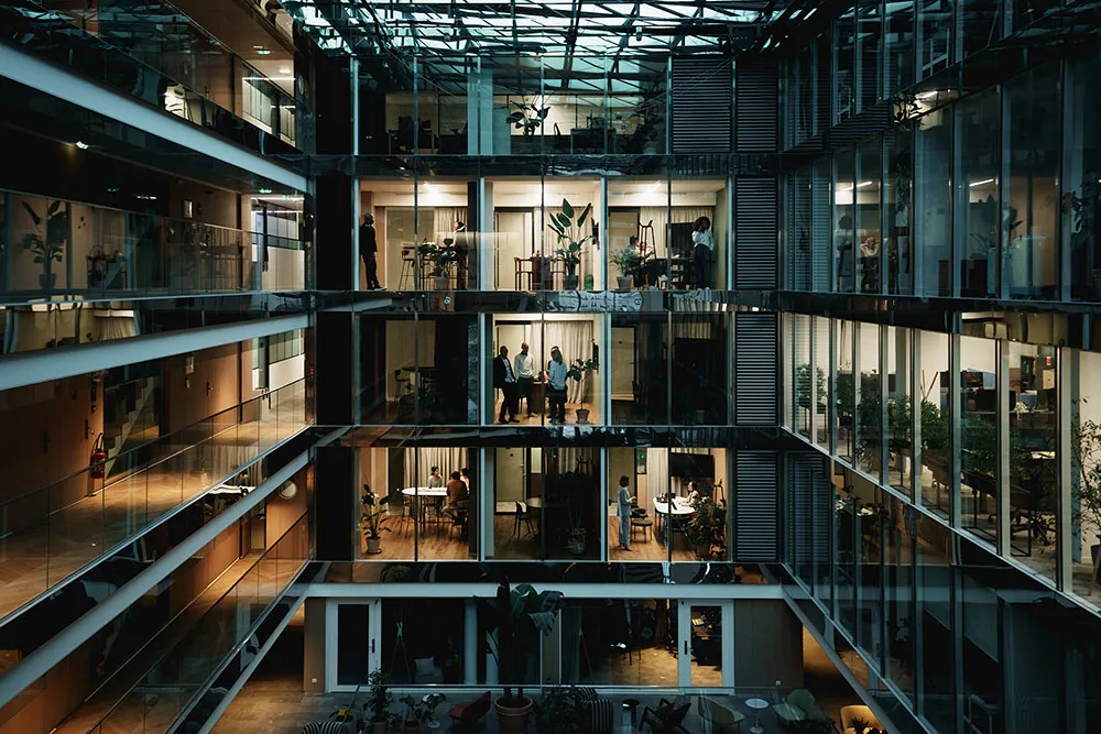 Et moderne kontorbygg med atrium i skumringen, med folk som jobber i glassrom over flere etasjer